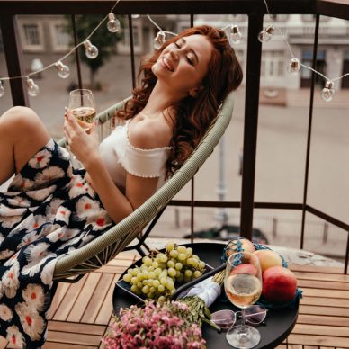 Young lady in floral skirt holds champagne glass and poses on terrace. Smiling girl sits next to table with fruits and white wine