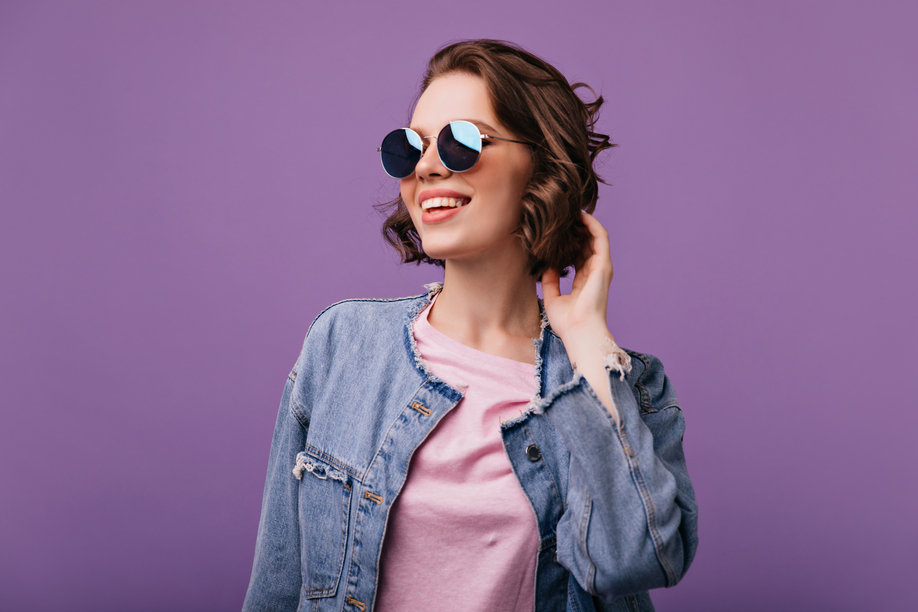 Appealing young woman in sparkle sunglasses looking in distance. Portrait of glamorous european model with short haircut smiling on purple background.