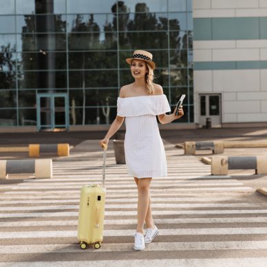 Attractive woman in white dress, hat and sneakers walks on crosswalk and smiles. Blonde girl holds passport and yellow suitcase.