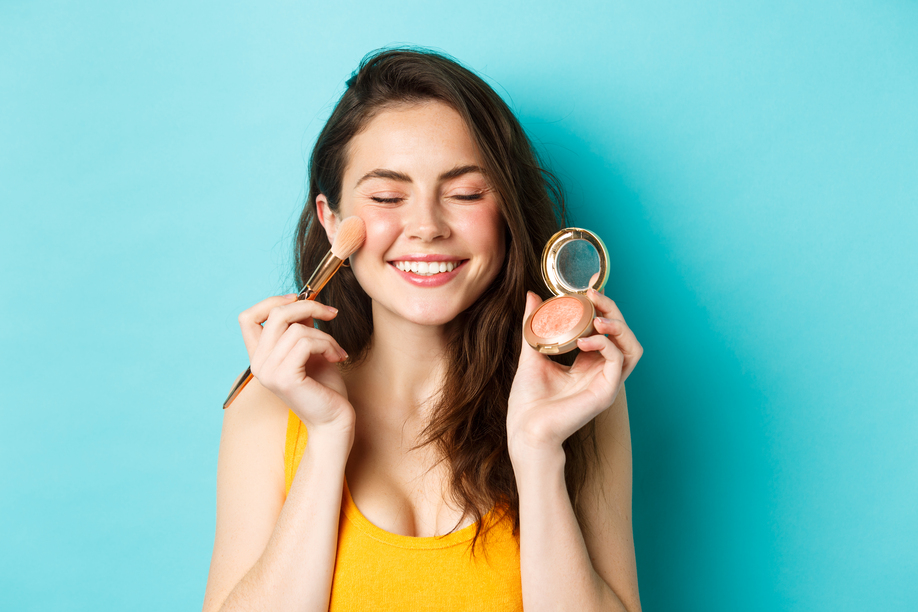 Beauty. Close up of beautiful young woman close eyes and smile while applying blushes on cheeks with make up brush, standing happy against blue background.