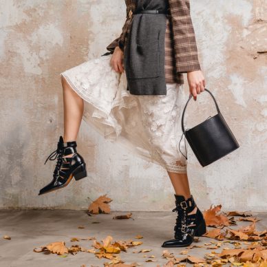 close up legs in boots of stylish woman in jacket coat walking against wall in street, autumn fashion trend, wearing white lace dress, handbag, leaves on the ground, urban style, footwear accessories