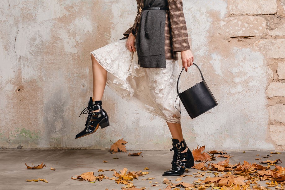 close up legs in boots of stylish woman in jacket coat walking against wall in street, autumn fashion trend, wearing white lace dress, handbag, leaves on the ground, urban style, footwear accessories