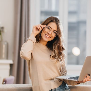 Cute girl in beige shirt touching glasses and holding laptop with smile. Indoor photo of beautiful female student preparing for lesson with computer at home.