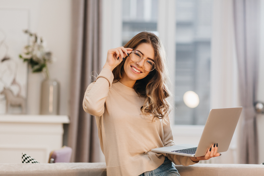 Cute girl in beige shirt touching glasses and holding laptop with smile. Indoor photo of beautiful female student preparing for lesson with computer at home.