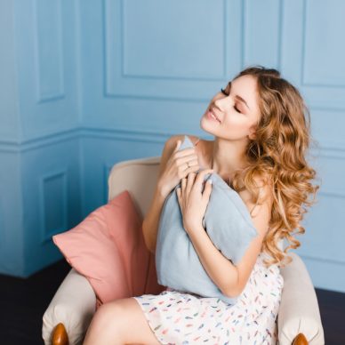 Cute girl with blond curly hair sits on armchair in studio with blue walls and holding a pillow. She wears white dress. She looks dreamy and beautiful.