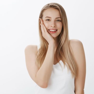 Tender young european girl with blond hair touching silk clear skin with palm and smiling sensually at camera as being satisfied with pores condition and facial color after applying skincare mask. Cosmetology, facial expressions and advertisement concept