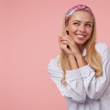 Indoor portrait of charming young woman with folded hands near her face, looking aside and smiling broadly, wearing casual clothes, posing over the pink background