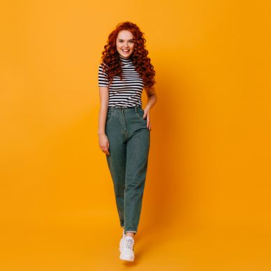 Lady in stylish denim pants and striped top posing on orange background. Full-length shot of redhead girl