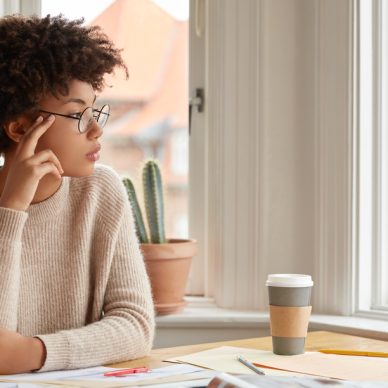 Photo of contemplative woman with Afro hairstyle, wears round spectacles, casual warm sweater, thinks about something while sits at desktop, drink coffee, poses near window indoor. Work concept