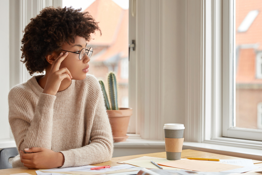 Photo of contemplative woman with Afro hairstyle, wears round spectacles, casual warm sweater, thinks about something while sits at desktop, drink coffee, poses near window indoor. Work concept