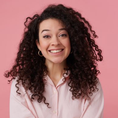 Portrait of attractive, adult girl with long dark curly hair. Wearing earrings and pastel pink shirt. Has make up. Broadly smiling. Watching at the camera isolated over pastel pink background