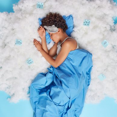 Relaxed curly haired Afro American woman sleeps well has sweet dreams being stress free wears sleepmask covered with blanket uses stickers for making necessary notes lies asleep on white cloud