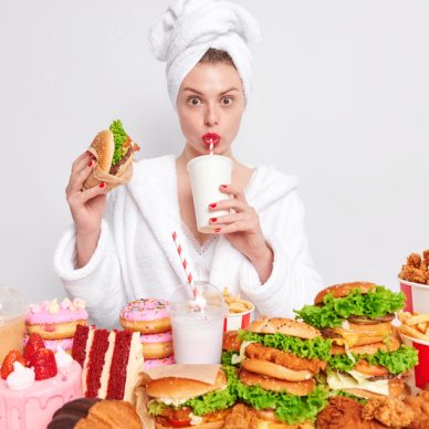 Unhealthy nuttrition concept. Housewife with red manicure and lips in domestic bathrobe towel on head drinks soda eats hamburger eats delicious fast food stands against white background. Breaking diet