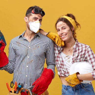 Professional male manual worker wearing protectibe eyewear on head, mask and gloves holding drilling machine fixing something and his colleague female with dirty face having happy expression