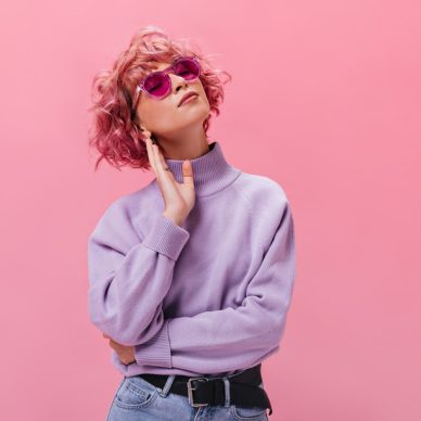 Attractive curly woman in purple cashmere sweater and fuchsia sunglasses poses on isolated background. Portrait of short-haired girl on pink backdrop .