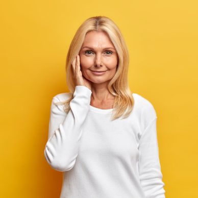 Attractive tender middle aged woman with blonde hair has healthy and wrinkled skin wears minimal makeup dressed in casual white jumper poses indoor against yellow background. Beauty age concept