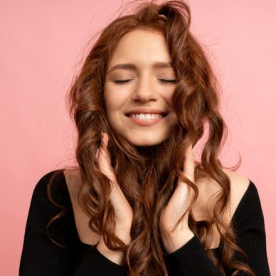 Beautiful red  head woman  with close eyes posing over pink background. Wavy hairs. Perfect smile.  Isolate in studio.