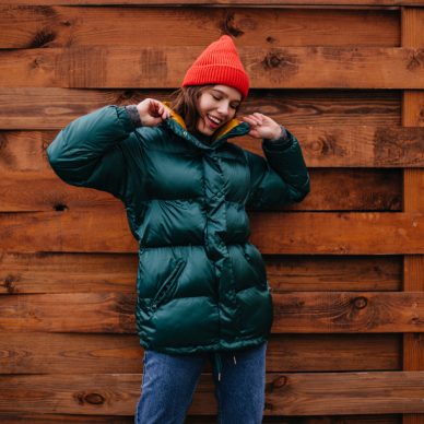 Fashionable girl in great mood posing on wooden background. Woman in baggy jeans, emerald coat and red hat smiling