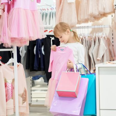 Pretty, cute girl holding shopping bags and choosing clothing in department store. Child looking at pink beautiful dress. Many pink clothes on hangers. Concept of stylish children clothing.