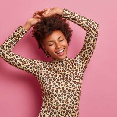 Happy dark skinned woman shakes body, raises hands and dances carefree, wears leopard jumper, closes eyes, enjoys awesome party, isolated over pink background, enjoys life, has playful mood.