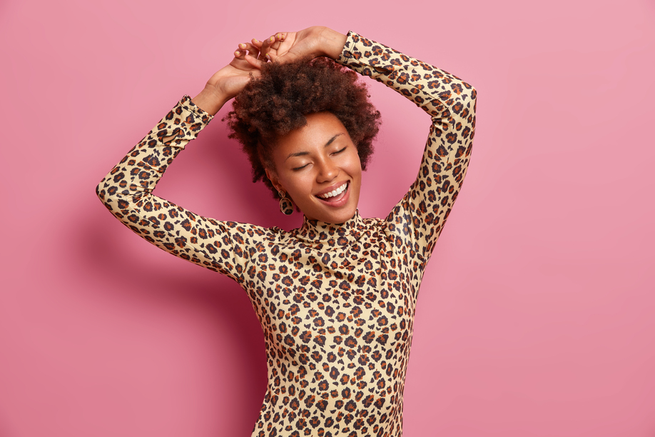 Happy dark skinned woman shakes body, raises hands and dances carefree, wears leopard jumper, closes eyes, enjoys awesome party, isolated over pink background, enjoys life, has playful mood.