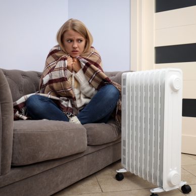 Heating season, woman sitting on sofa and warm up with heater