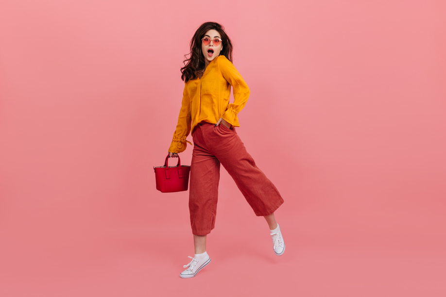 Perky girl in stylish glasses stares into camera in amazement, walking on pink background. Brunette in culottes and orange blouse posing with red handbag