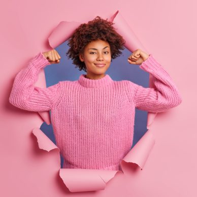 Self assured beautiful young Afro American woman raises arms shows biceps being strong and powerful proud of her own achievements wears casual knitted sweater breaks through paper background