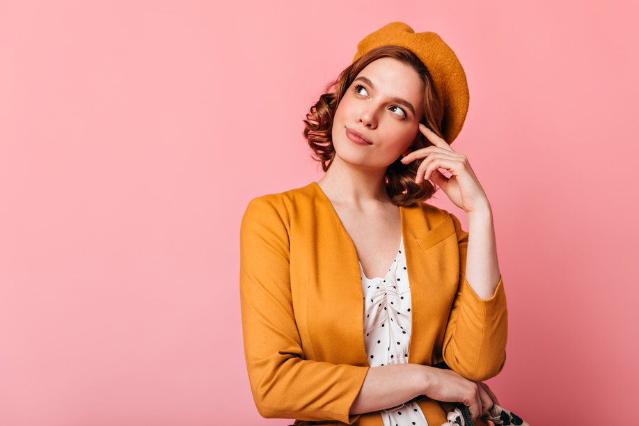Studio shot of pensive french girl looking up. Charming young woman in beret thinking about something on pink background.