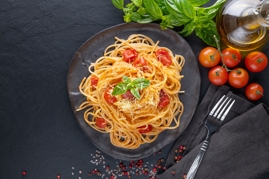 Tasty appetizing classic italian spaghetti pasta with tomato sauce, cheese parmesan and basil on plate  and ingredients for cooking pasta on dark table. Flat lay top view copy spce.