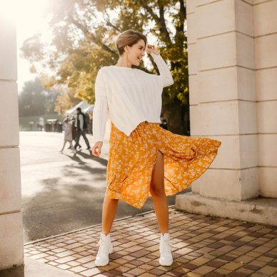 young pretty happy smiling woman in yellow printed dress and knitted white sweater on sunny autumn day having fun in street wearing stylish outfit and white boots