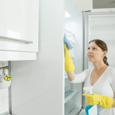 Young worker woman wearing rubber protective yellow gloves cleaning the fridge with rag and spray bottle detergent. Home, housekeeping concept. Lifestyle