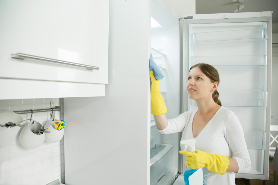 Young worker woman wearing rubber protective yellow gloves cleaning the fridge with rag and spray bottle detergent. Home, housekeeping concept. Lifestyle