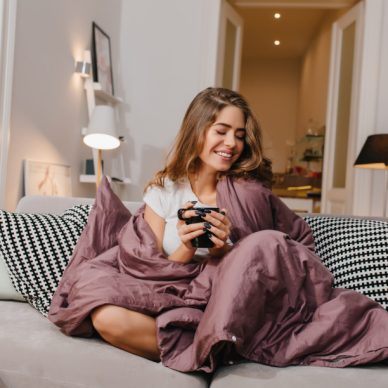 Cheerful girl sitting on couch with blanket and cushions and smiling. Spectacular brunette lady laughing, while drinking coffee under plaid.