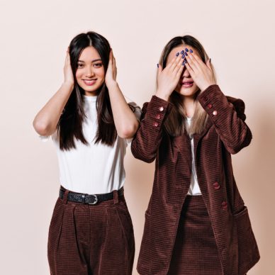 Dark-haired girl smiles and covers her ears, blonde woman in corduroy suit covers her eyes with her hands