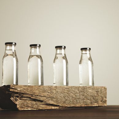 Four clear transparent glass bottles with black caps filled with pure drinking water presented on wooden brick, isolated on white