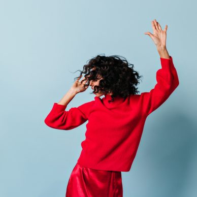 Glad brunette curly woman dancing on blue background. Studio shot of stunning female model in red outfit.