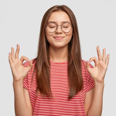 Pleased satisfied young female model makes zero gesture, wears transparent glasses, has long dark hair, dressed in casual striped t shirt, stands over white background. People, body language concept