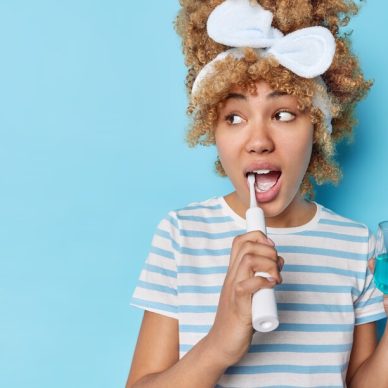 Photo of blonde curly woman brushes teeth with electric brush holds glass of mouthwash for fresh breath isolated over blue background blank space for your promotional content. Dental care concept