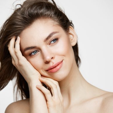 Beautiful naked young girl with perfect clean skin smiling touching hair over white wall. Facial treatment. Copy space.
