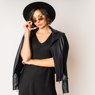 Fashionable model in black hat, evening dress and leather jacket posing on white background in studio.