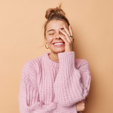 Happy emotions and feelings concept. Joyful young woman with combed hair makes face palm smiles toothily wears knitted sweater isolated over beige background laughs at something funny feels glad