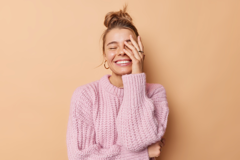 Happy emotions and feelings concept. Joyful young woman with combed hair makes face palm smiles toothily wears knitted sweater isolated over beige background laughs at something funny feels glad