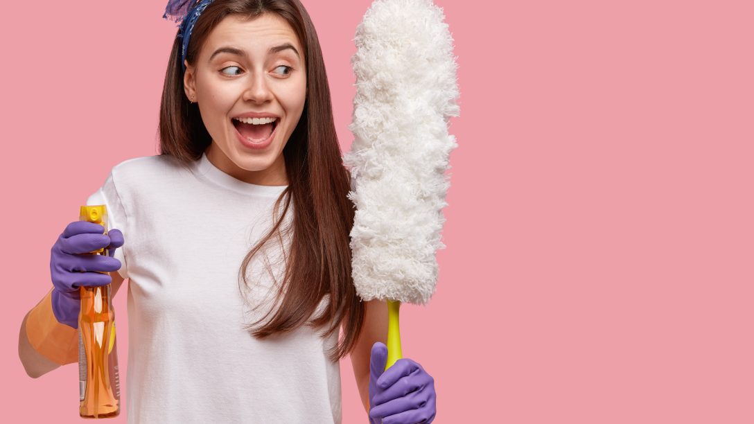 Positive European woman wears gloves and headband, looks joyfully aside, holds spray and brush for cleaning house, has busy weekends, models over pink studio wall with copy space for information