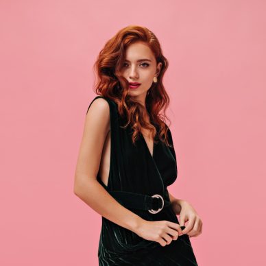 Attractive woman in dark dress looks into camera on pink background. Curly ginger girl with rose lipstick in modern green clothes posing..