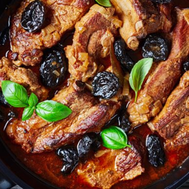 Stewed short pork ribs with dried plums or prunes, pork broth, onion, and garlic, decorated with fresh basil, served on a black baking dish on an old barn wood background, close-up