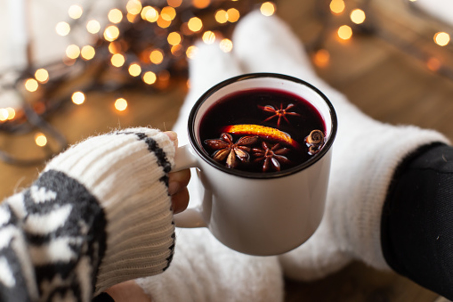 Composition with a mug of hot red wine mulled wine with spices. Cozy holiday atmosphere, plush socks, warm sweater, Christmas lights on a dark background.