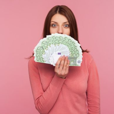 Amazed happy woman with brown hair in pink sweater hiding face behind fan of euro banknotes, interest-free cash withdrawal. Indoor studio shot isolated on pink background