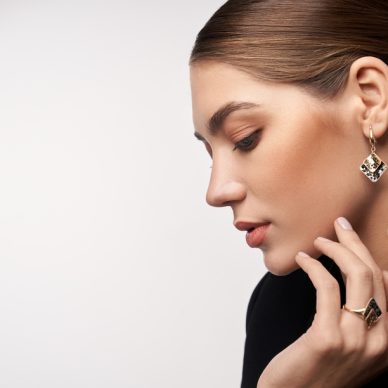 Close up portrait of young beautiful brunette female model presenting golden and silver earrings and ring. Side view of woman in formal suit posing in studio, isolated on white background.