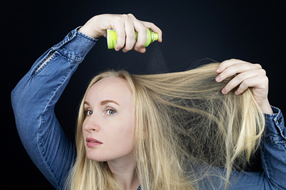 Blonde girl sprays shampoo on her hair. The problem of oily hair while traveling. An emergency remedy for excessive sebum production. Make your head clean without water. Dry shampoo.